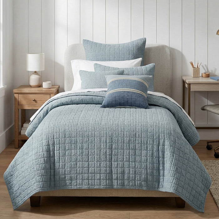Blue textured quilt and pillows on a bed in a bedroom setting.