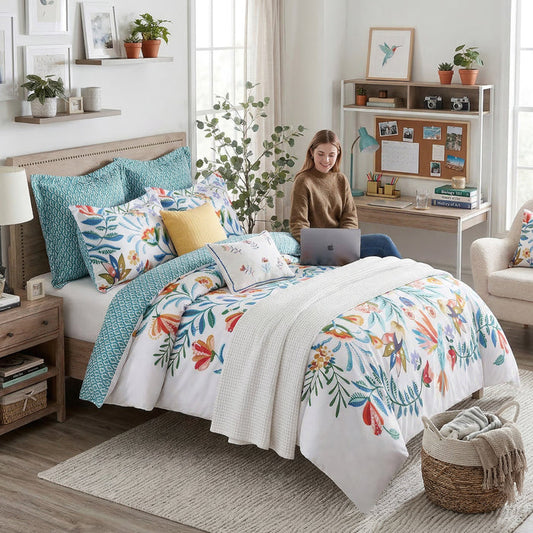 Woman using a laptop on a bed with colorful bedding in a bright bedroom.