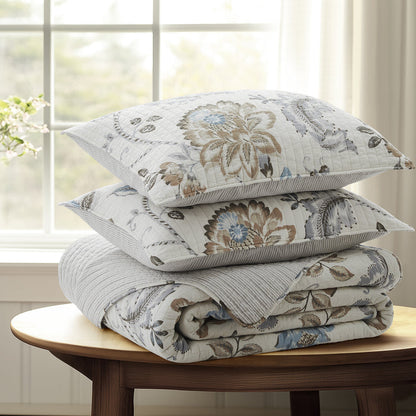 Stack of floral-patterned pillows and blanket on a wooden table with a window in the background